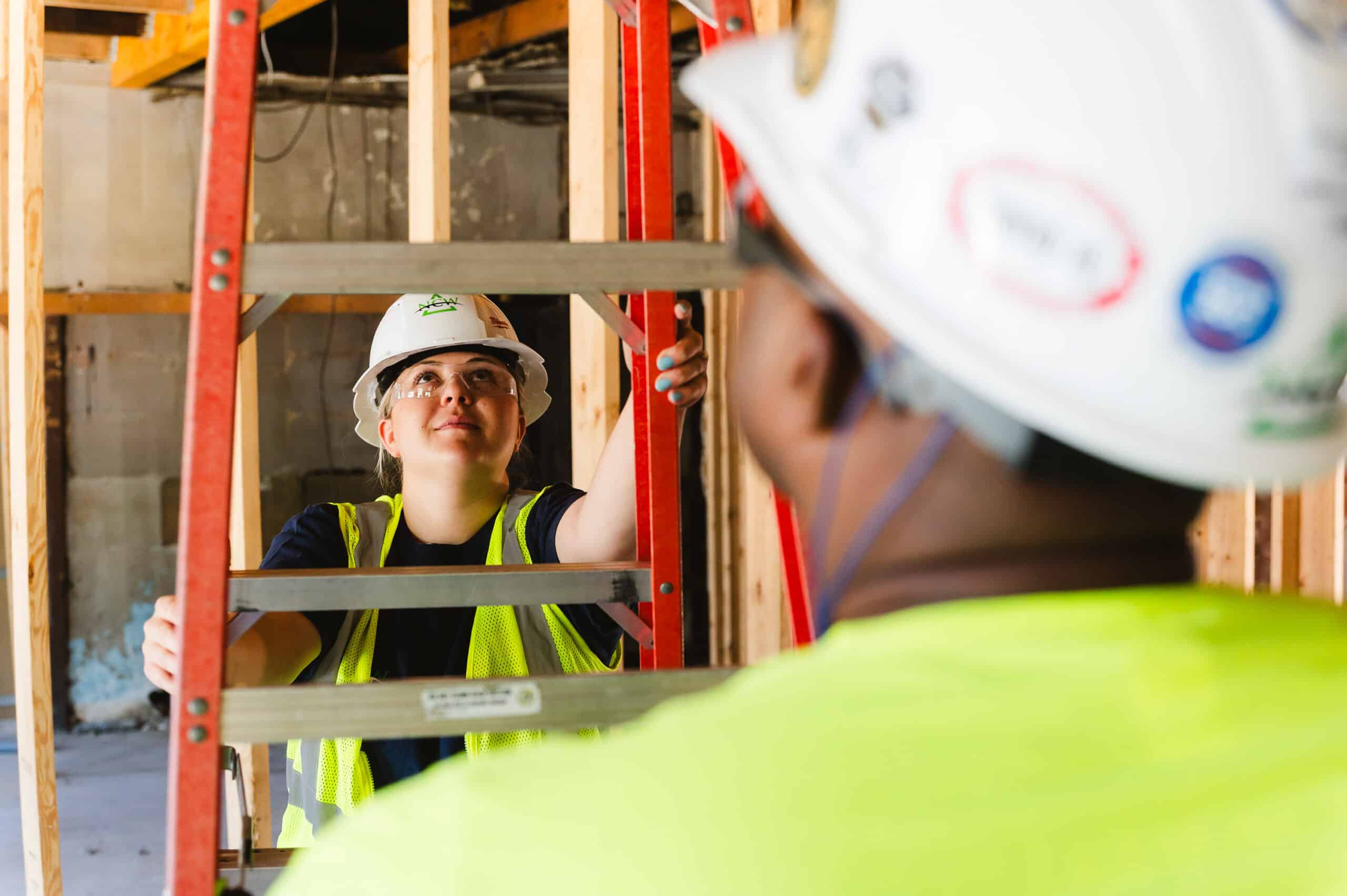 A construction worker climbs a ladder with a spotter while wearing personal protective equipment