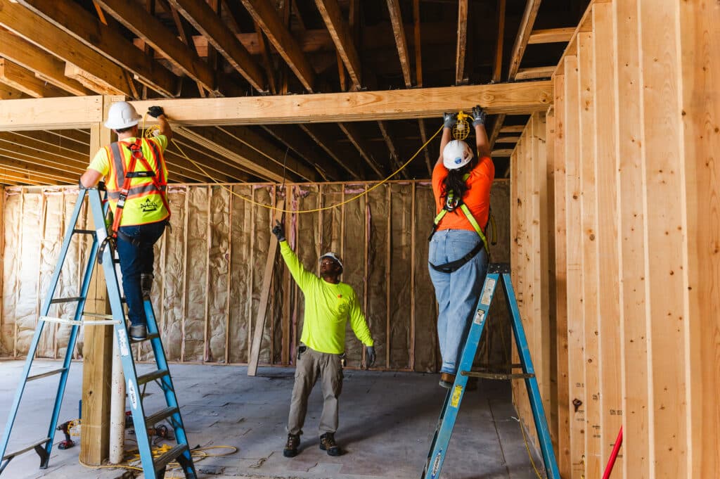 Three construction workers hang a string of work lights on a job site using ladders and personal protective equipment.  